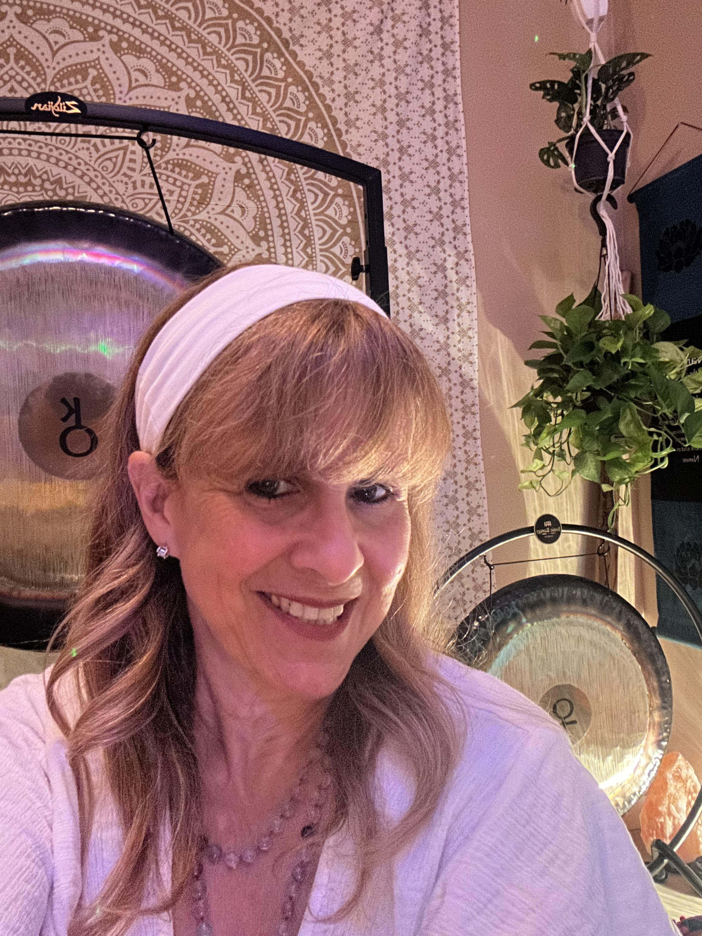 Diana smiling in white with a headband beside two Paiste planetary gongs under hanging plants and a mandala tapestry